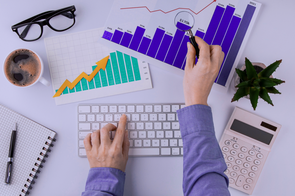 A top-down photo of a desk with a person typing on a keyboard and reaching over to a blue printed graph.