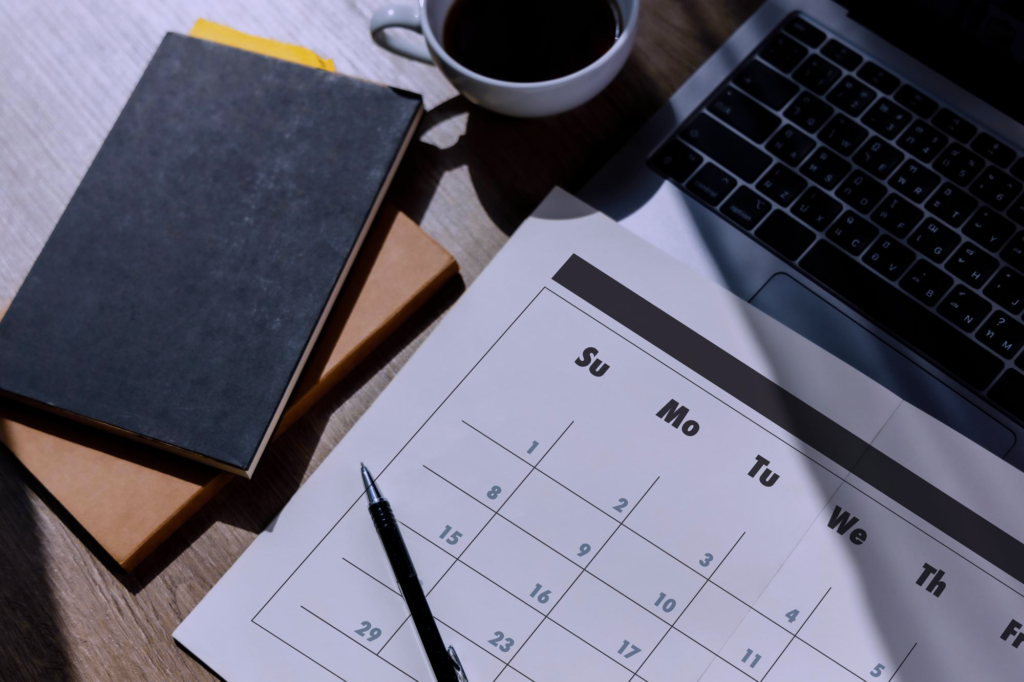 Um calendário de papel, alguns cadernos, uma xícara de café e um laptop colocados um ao lado do outro sobre uma mesa de madeira.