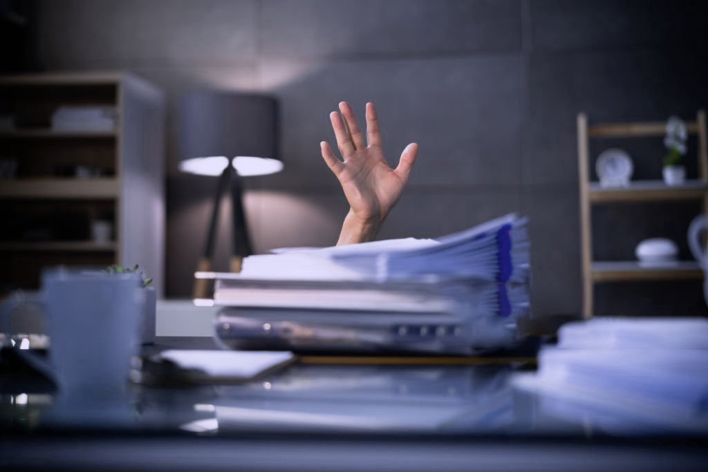 A high depth of field image of a person's raised hand over a pile of papers, signifying work overload and the feeling of overworking.