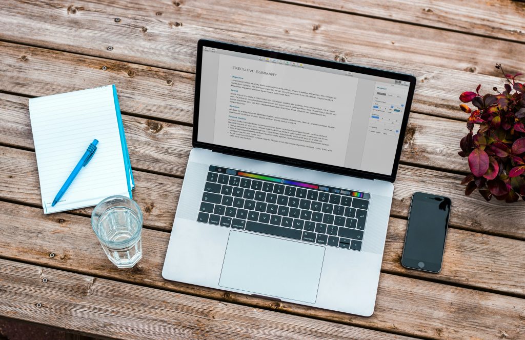 Um laptop, um telefone e um caderno de papel sobre uma mesa de madeira, simbolizando multitarefa e produtividade.