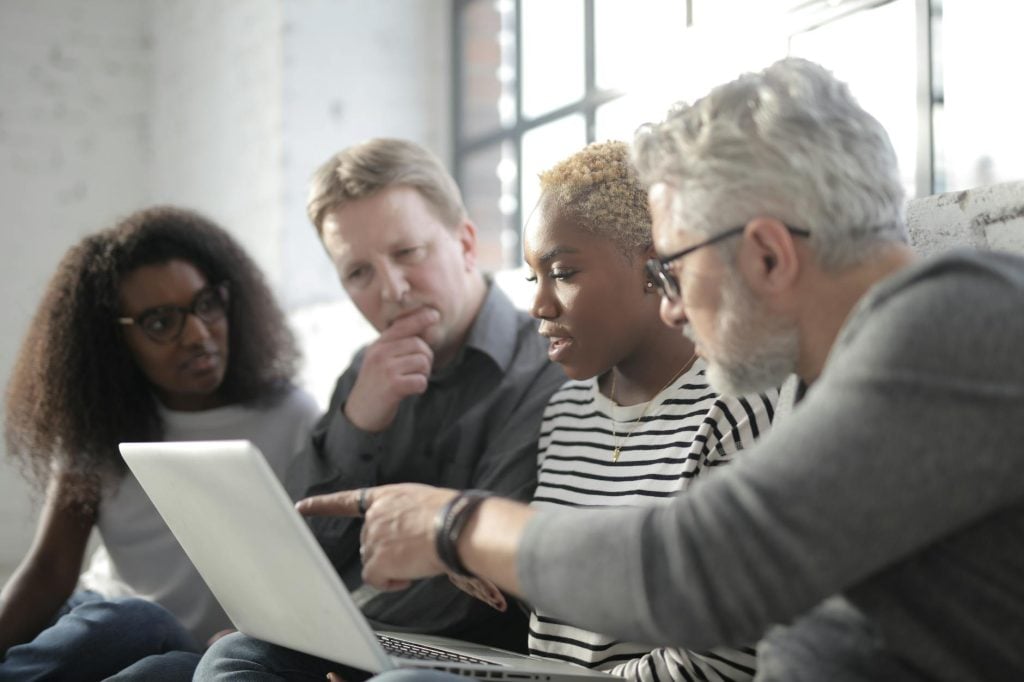 Middle aged employee in eyeglasses pointing at netbook screen with finger while sitting near multiracial pensive colleagues near window in afternoon