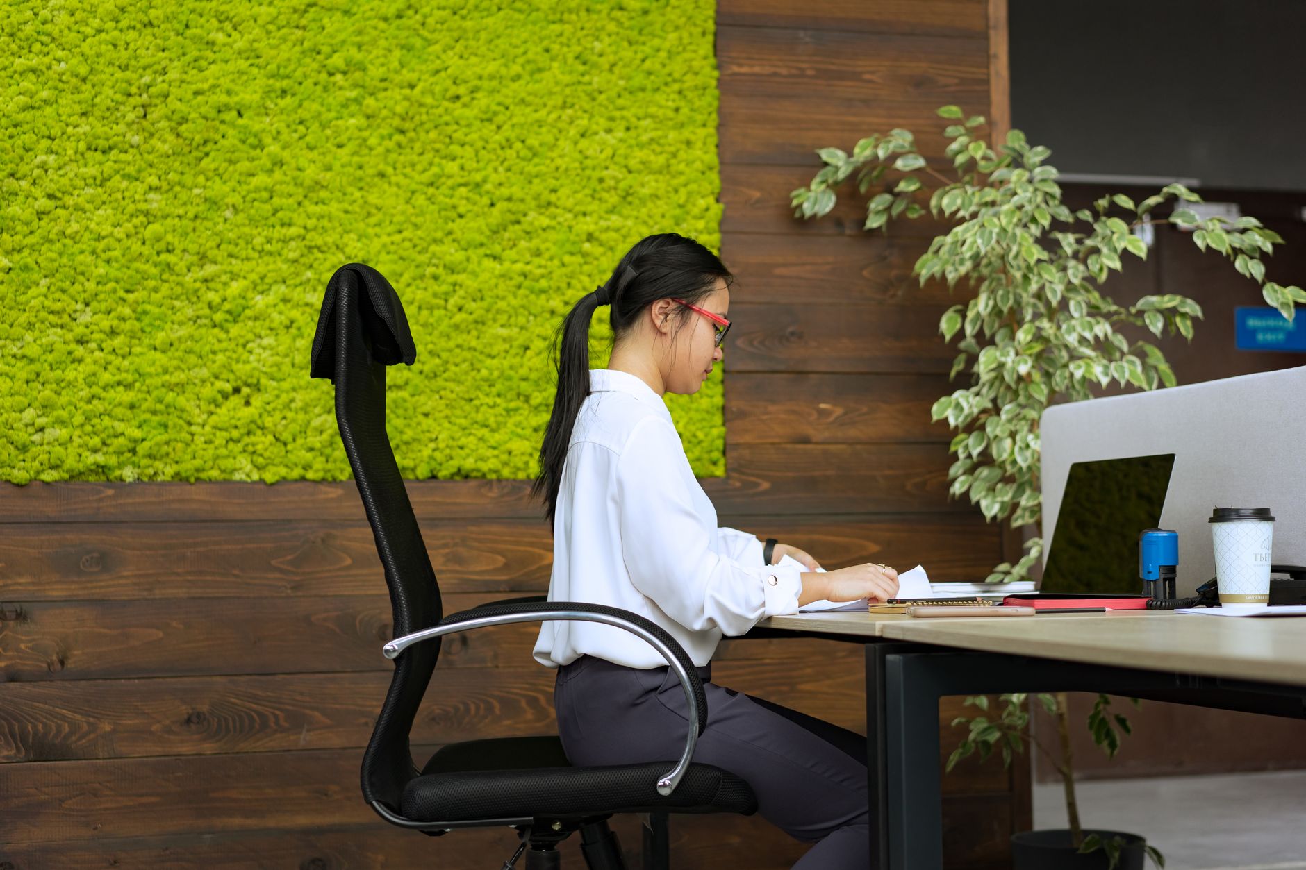 Woman working at her desk in a modern office with green moss wall and plants.
