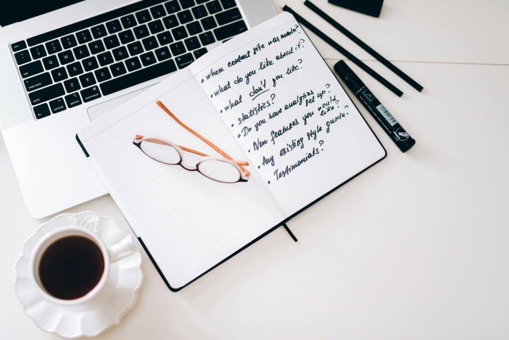 A stylish workspace featuring a laptop, notebook with notes, glasses, and a coffee cup.